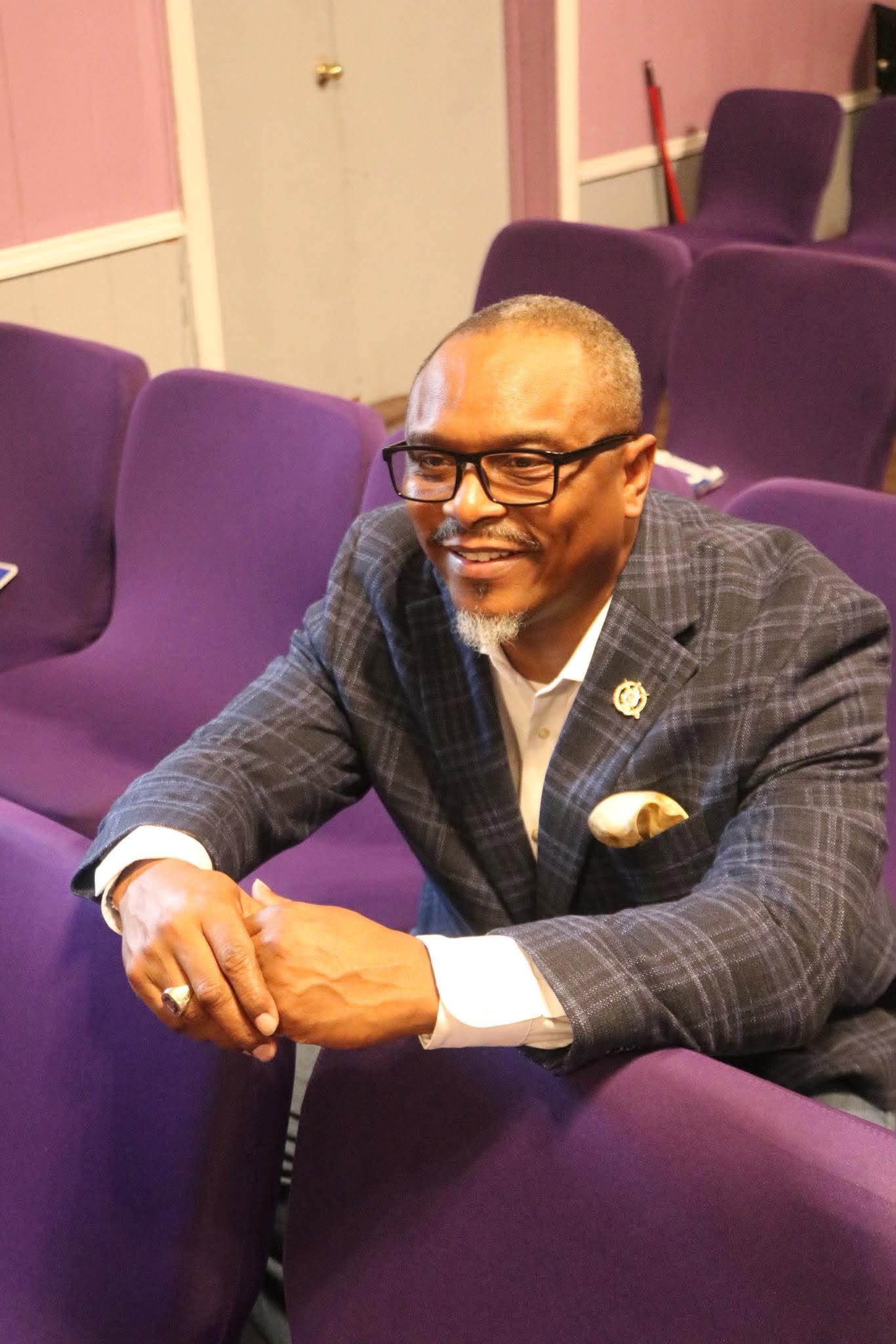 Richard Wooten smiling and leaning forward among purple church chairs.